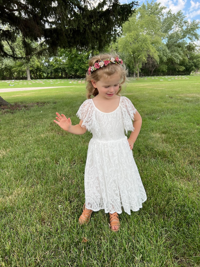 May include: A young girl wearing a white lace dress with a floral crown. She is standing in a grassy area with trees in the background.