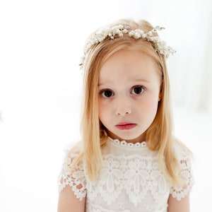 May include: A young girl with blonde hair wearing a white lace dress and a pearl and silver headband. She is looking at the camera with a serious expression.