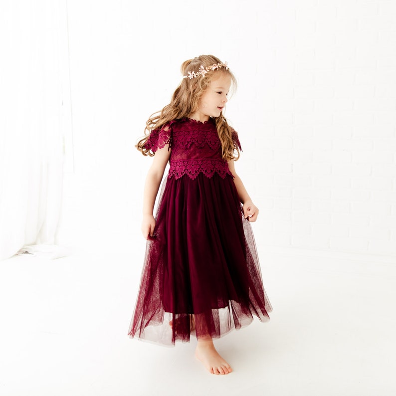 May include: A young girl wearing a burgundy tulle dress with a lace bodice and a gold headband. The dress has a full skirt and is perfect for a special occasion.