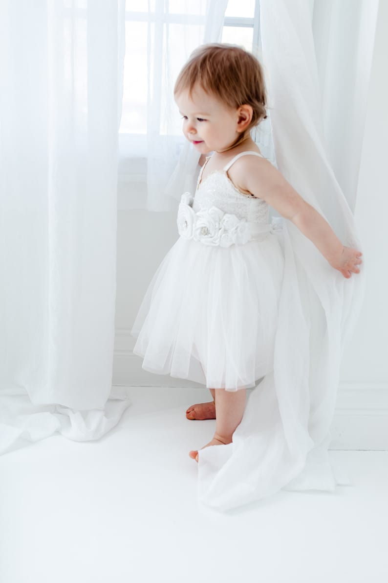 Puede incluir: Una ni&ntilde;a peque&ntilde;a con un vestido blanco de tul con un adorno floral alrededor de la cintura se para frente a una cortina blanca. Ella est&aacute; mirando hacia la izquierda del marco.