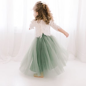 May include: A young girl wearing a white lace top and a sage green tulle skirt. She is standing in front of a white window with white curtains.