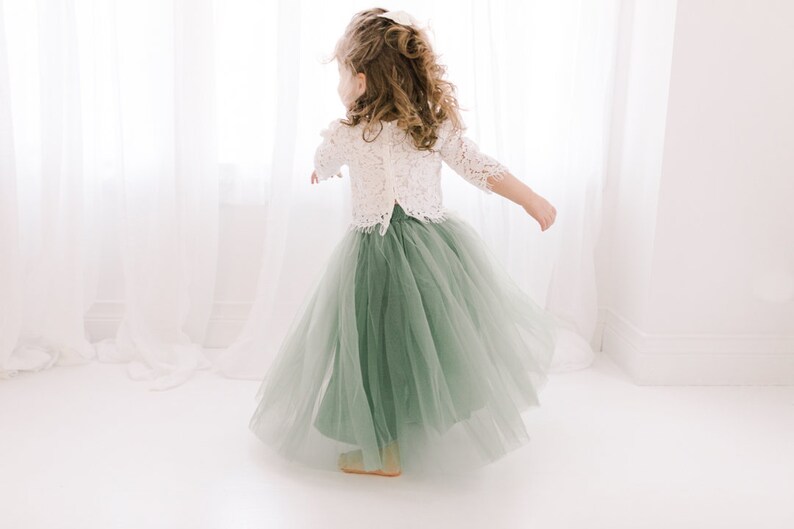 May include: A young girl wearing a white lace top and a sage green tulle skirt. She is standing in front of a white window with white curtains.