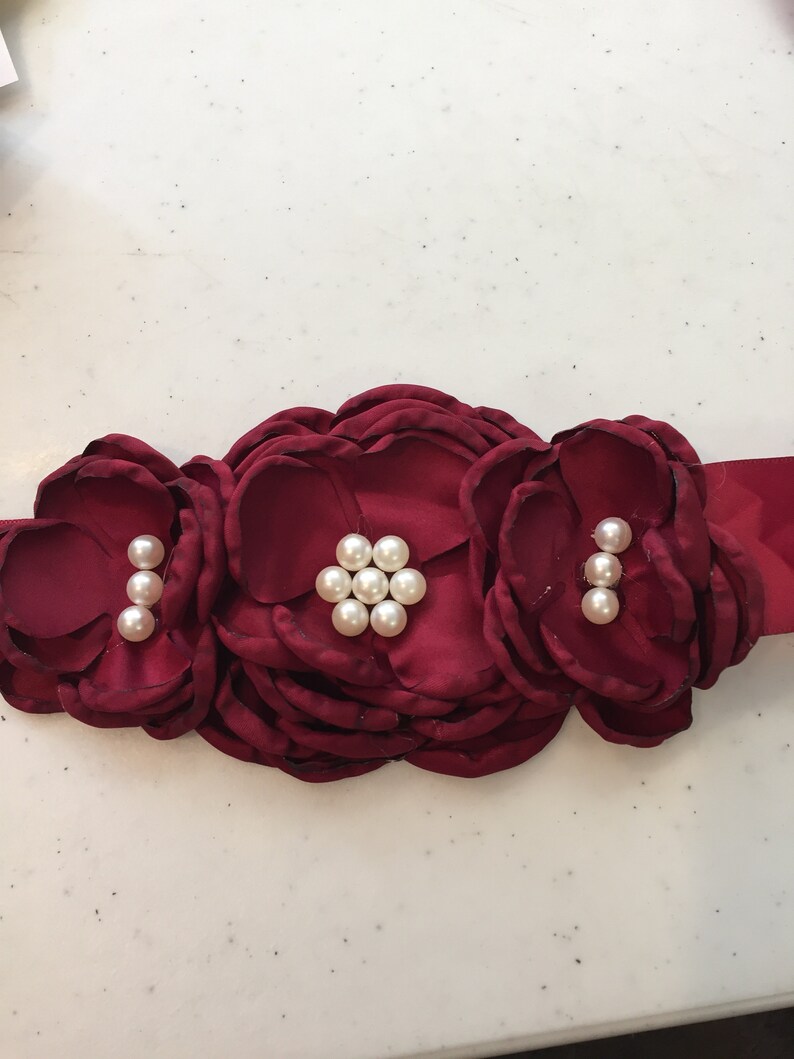 May include: A burgundy satin sash with three large fabric flowers, each adorned with pearl-like embellishments.