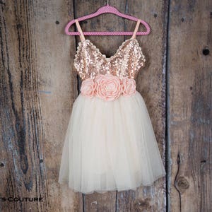 May include: A tulle dress with a rose gold sequin bodice and a peach flower sash. The dress is hanging on a pink polka dot hanger against a wooden background.