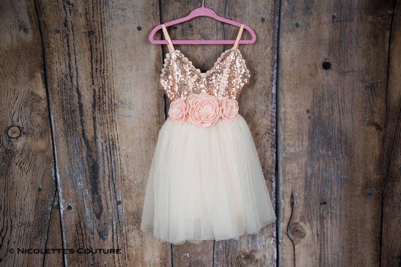 May include: A tulle dress with a rose gold sequin bodice and a peach flower sash. The dress is hanging on a pink polka dot hanger against a wooden background.