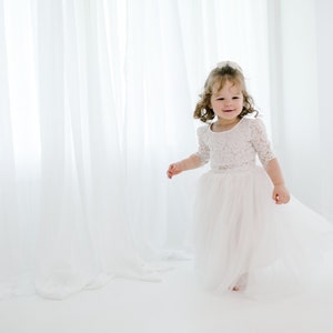 May include: A young girl in a white lace dress with a tulle skirt smiles and twirls in a white room.