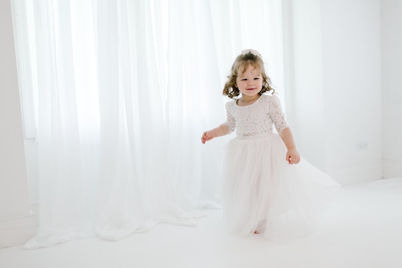 May include: A young girl in a white lace dress with a tulle skirt smiles and twirls in a white room.