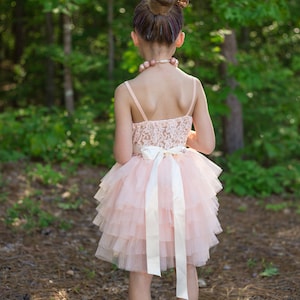 May include: A young girl wearing a pink tulle dress with a white satin bow and a pearl necklace. She is standing in a wooded area with green trees and brown ground.