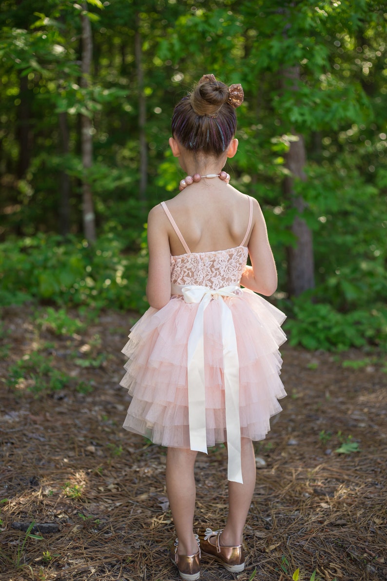 May include: A young girl wearing a pink tulle dress with a white satin bow and a pearl necklace. She is standing in a wooded area with green trees and brown ground.