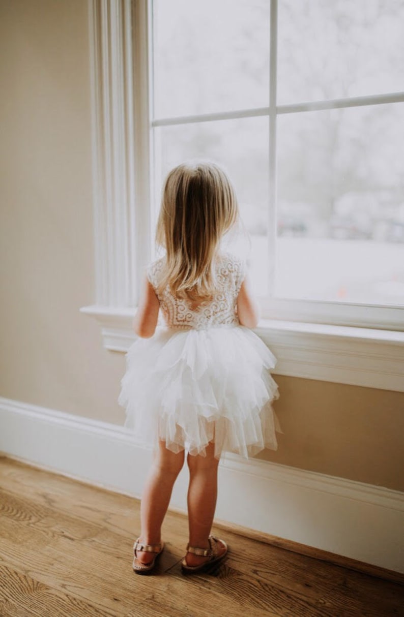 May include: A young girl in a white tulle dress with lace detailing stands in front of a window, looking out. She is wearing brown sandals.