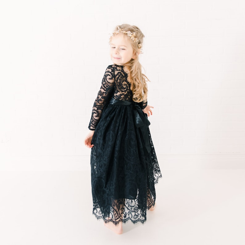 May include: A black long-sleeved dress featuring an intricate lace overlay with a floral pattern, a scalloped hemline, and a solid black satin ribbon tied in a bow at the waist. The dress has a fitted bodice and a full, flowing skirt.