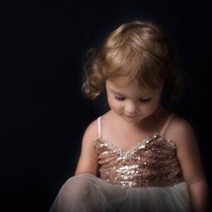 May include: A young girl with blonde curly hair wearing a sparkly gold dress and a white tulle skirt. She is looking down at something off camera.