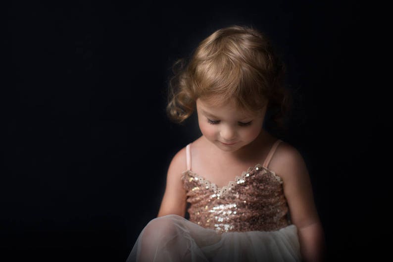 May include: A young girl with blonde curly hair wearing a sparkly gold dress and a white tulle skirt. She is looking down at something off camera.