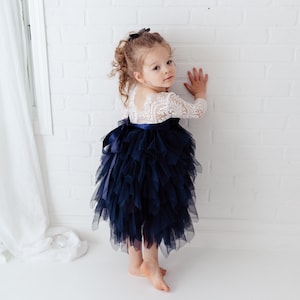 May include: A dress featuring a white lace bodice with three-quarter length sleeves and a voluminous navy blue tulle skirt. The skirt is layered with ruffled fabric and accented by a dark blue satin ribbon tied in a bow at the waist.