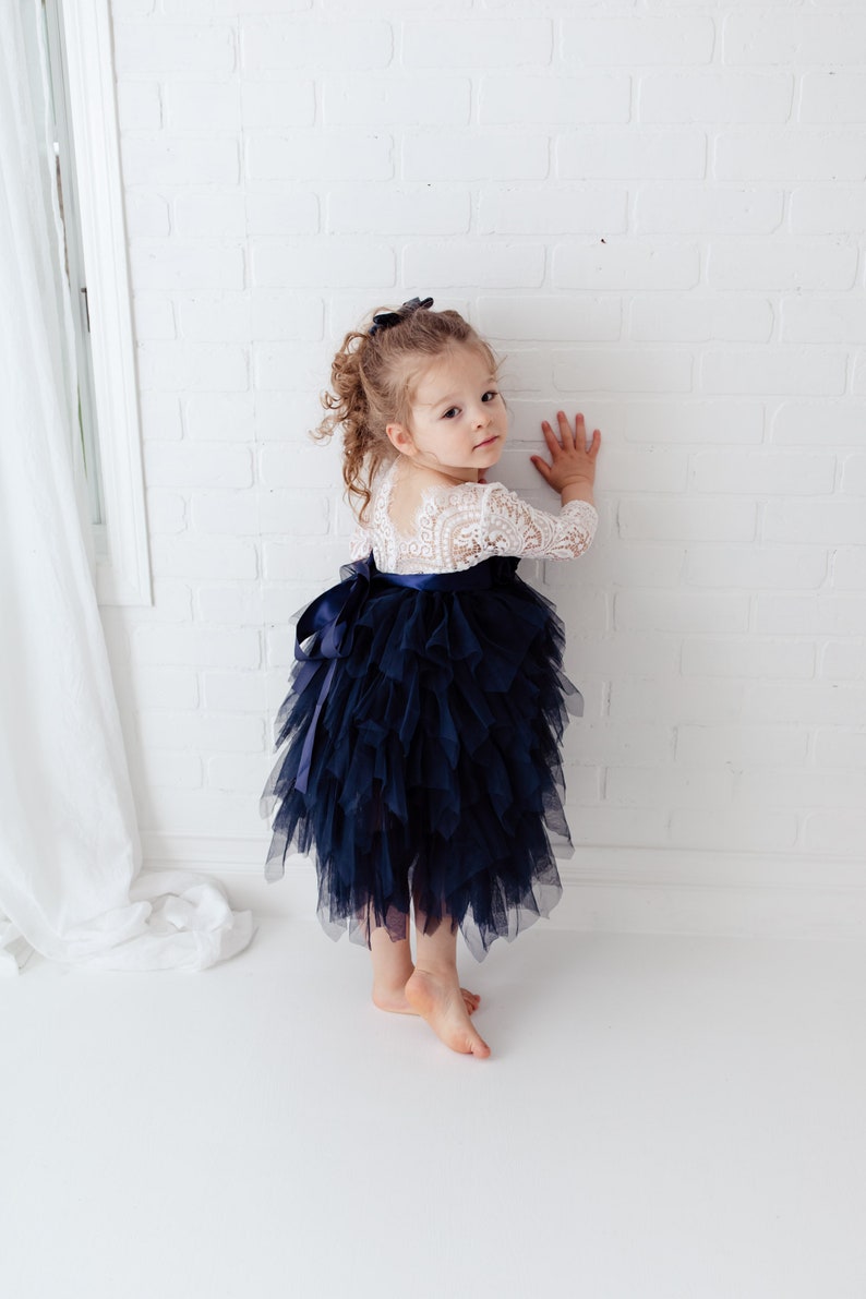 May include: A dress featuring a white lace bodice with three-quarter length sleeves and a voluminous navy blue tulle skirt. The skirt is layered with ruffled fabric and accented by a dark blue satin ribbon tied in a bow at the waist.