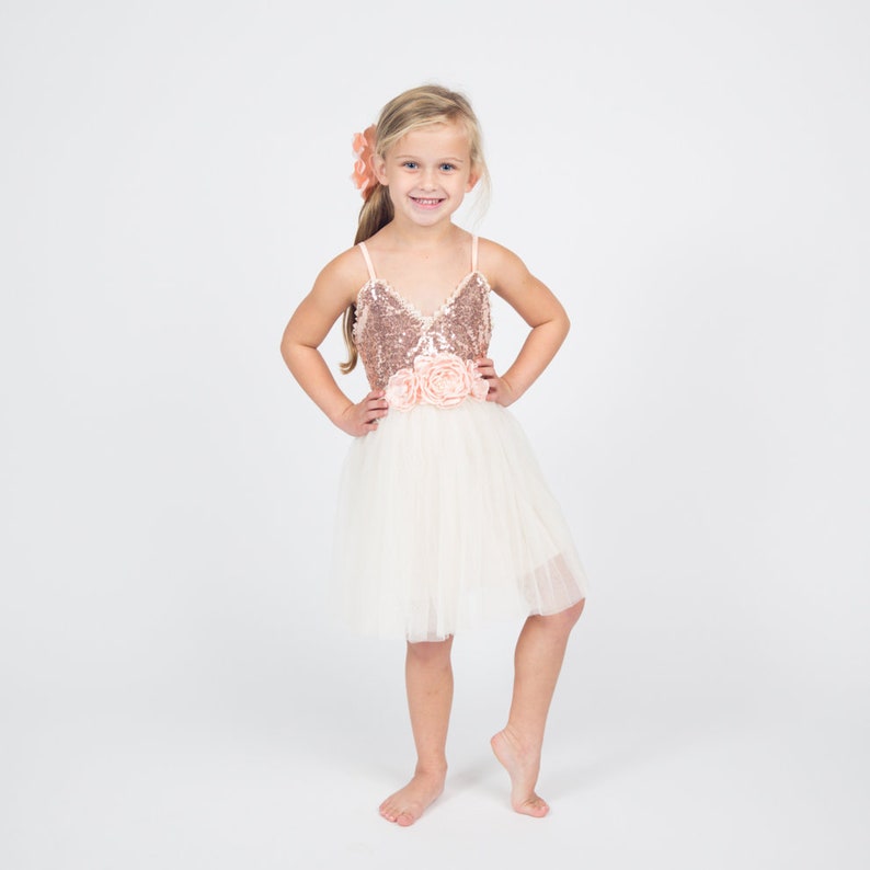 May include: A young girl wearing a white tulle dress with a rose gold sequined bodice and a pink flower sash. She is smiling and has her hair pulled back with a pink flower clip.