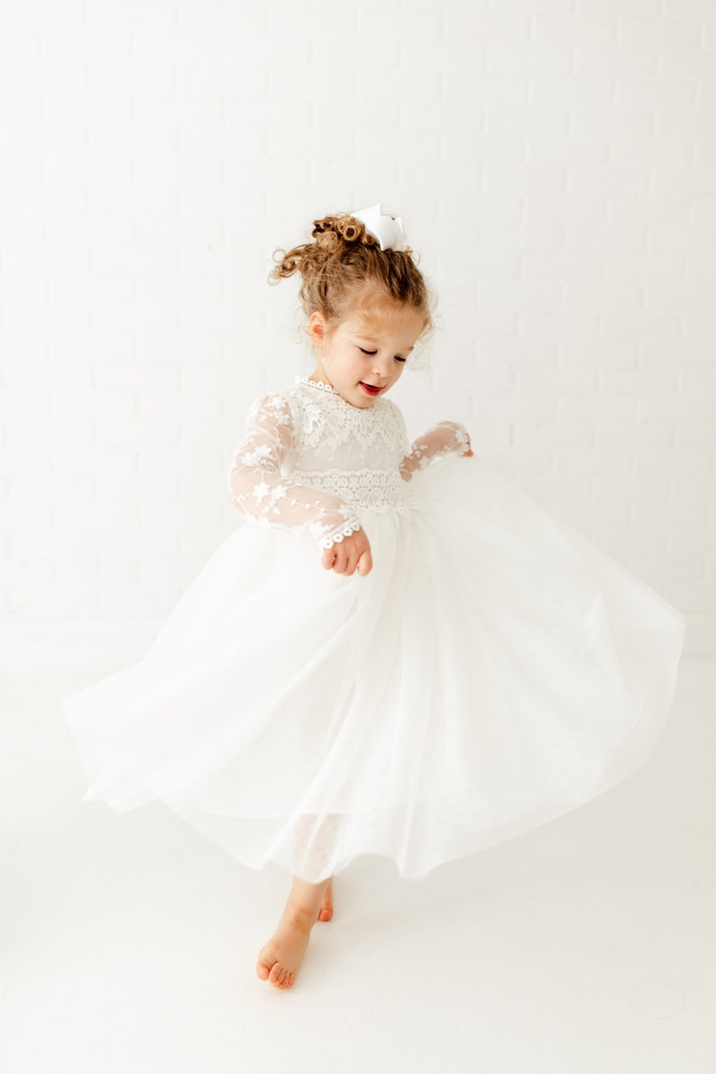 May include: A young girl wearing a white tulle dress with lace detailing and long sleeves. She is twirling and smiling.