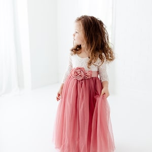 May include: A young girl wearing a white lace top with a pink tulle skirt and a large pink flower on the waist.