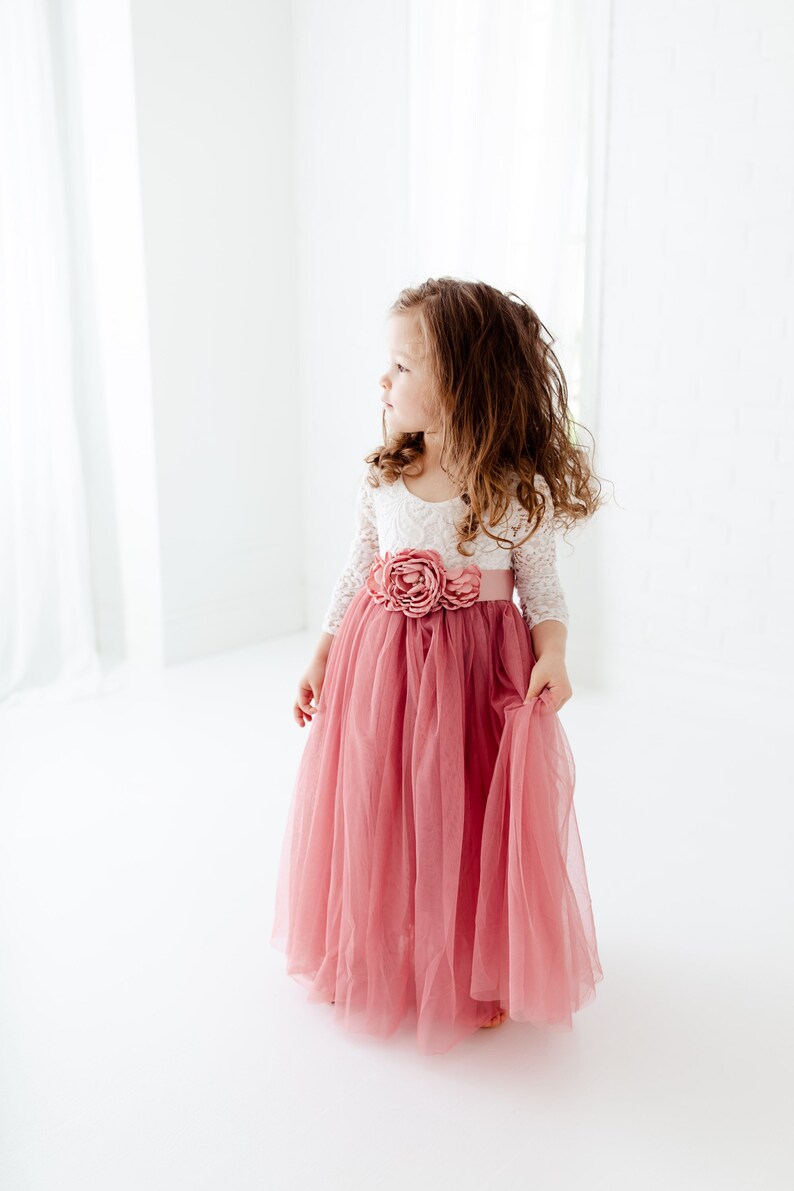 May include: A young girl wearing a white lace top with a pink tulle skirt and a large pink flower on the waist.