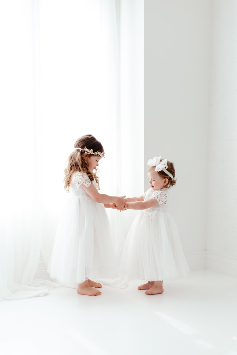 Op de afbeelding: Twee kinderen in witte jurken met kanten mouwtjes en tule rokken. Het langere kind draagt een goudkleurige bloemenhaarband, terwijl het kleinere kind een witte strikhaarband draagt. Beide jurken hebben lijfjes van gedetailleerd kant.