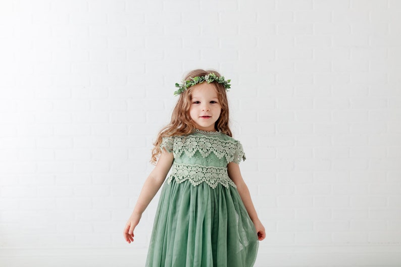 May include: A young girl wearing a sage green tulle dress with lace detailing and a green leaf crown. The dress has a round neckline and short sleeves.