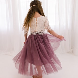 May include: A young girl wearing a white lace top with a button-up back and a mauve tulle skirt. The skirt is flowing and the girl is twirling.