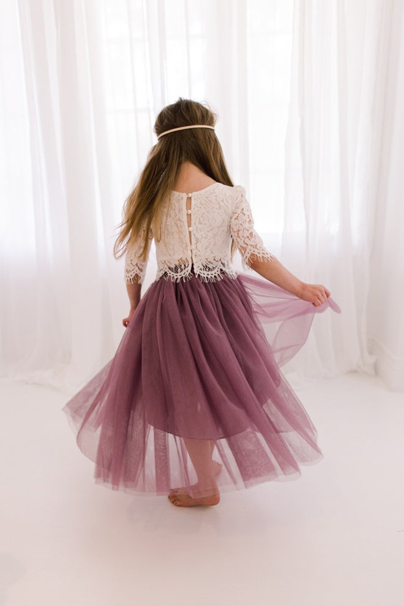 May include: A young girl wearing a white lace top with a button-up back and a mauve tulle skirt. The skirt is flowing and the girl is twirling.