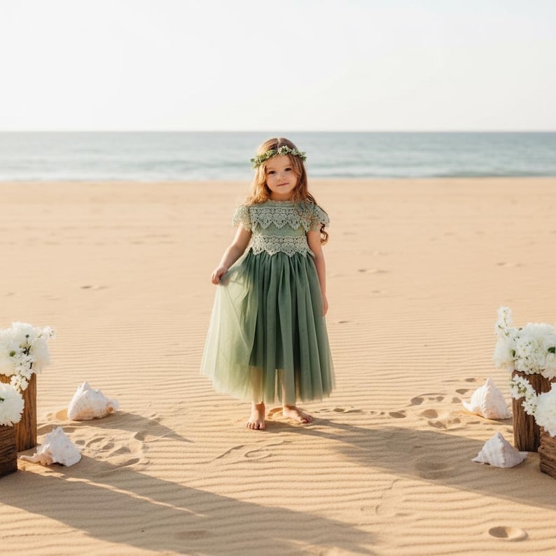 May include: A sage green dress featuring a lace bodice with short sleeves and a layered tulle skirt that falls to the mid-calf. The dress is paired with a matching floral crown made of small green leaves, worn on the head of the individual.