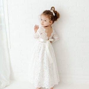 May include: A floor-length white lace dress with long sleeves, a scoop back, and a wide satin ribbon tied in a bow at the waist. The dress features intricate floral lace patterns throughout the bodice and skirt, with a scalloped hemline.