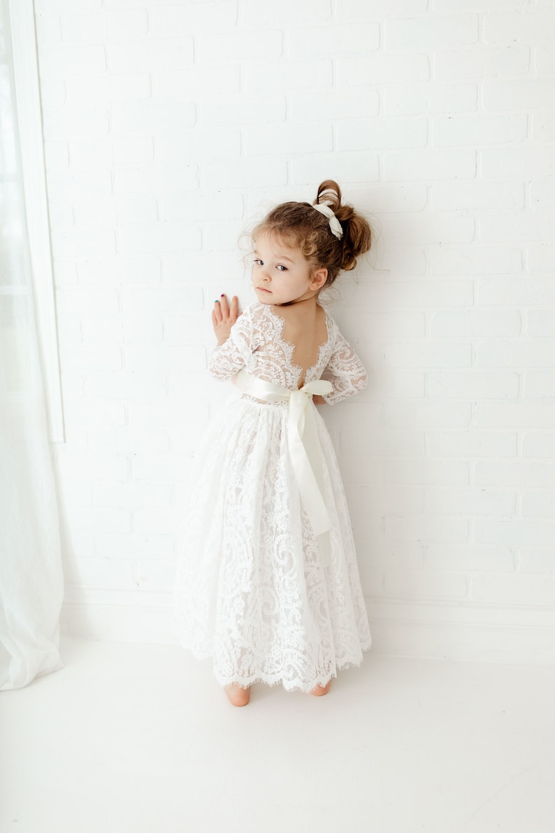 May include: A floor-length white lace dress with long sleeves, a scoop back, and a wide satin ribbon tied in a bow at the waist. The dress features intricate floral lace patterns throughout the bodice and skirt, with a scalloped hemline.