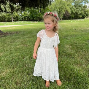 May include: A young girl wearing a white lace dress with a floral headband. She is standing in a grassy area with trees in the background.