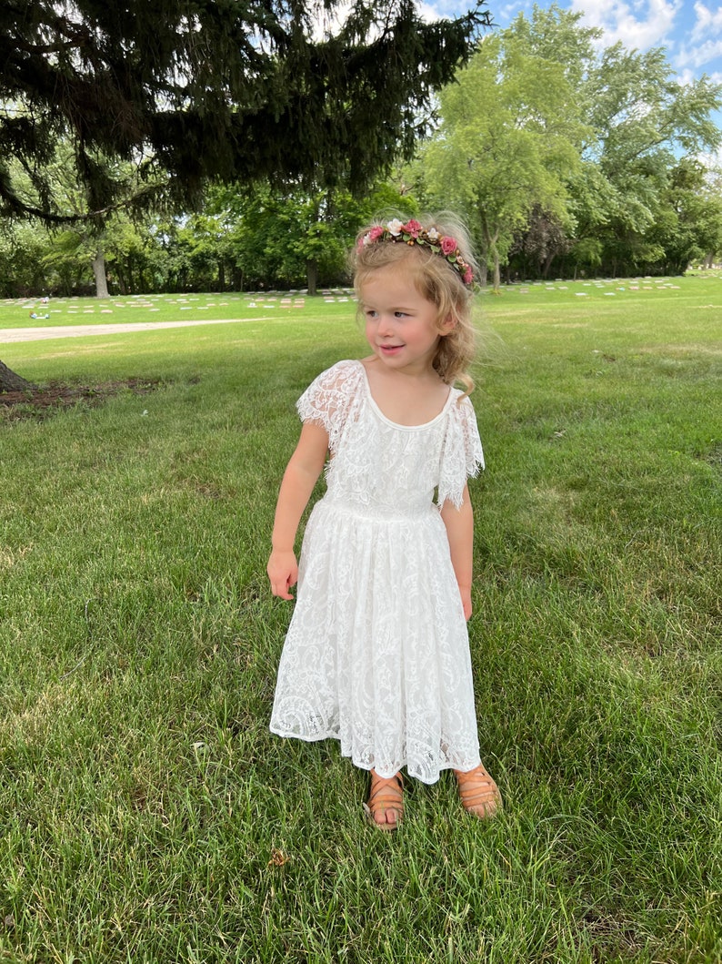 May include: A young girl wearing a white lace dress with a floral headband. She is standing in a grassy area with trees in the background.