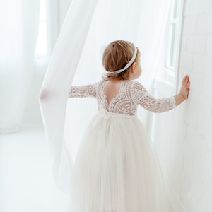 Pu&ograve; includere: Una bambina che indossa un abito bianco di pizzo con una gonna di tulle. &Egrave; in piedi in una stanza bianca con una tenda bianca dietro di lei.