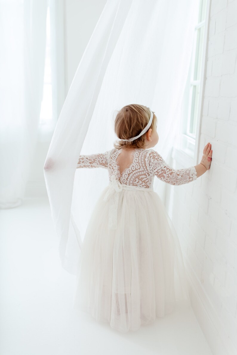 Pu&ograve; includere: Una bambina che indossa un abito bianco di pizzo con una gonna di tulle. &Egrave; in piedi in una stanza bianca con una tenda bianca dietro di lei.