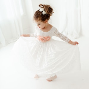 May include: A white lace dress with long sleeves and a scoop neckline, featuring a waistline adorned with three large, soft peach-colored fabric flowers. The skirt is full and flared, and the hair is styled in an updo with a white bow accessory.