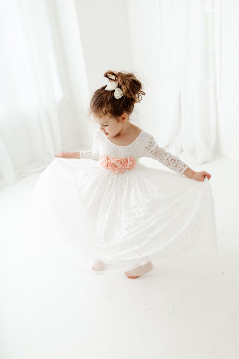 May include: A white lace dress with long sleeves and a scoop neckline, featuring a waistline adorned with three large, soft peach-colored fabric flowers. The skirt is full and flared, and the hair is styled in an updo with a white bow accessory.