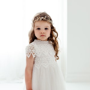May include: A young girl wearing a white lace dress with a gold and white flower crown. She has long brown hair and is looking directly at the camera.