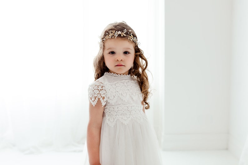 May include: A young girl wearing a white lace dress with a gold and white flower crown. She has long brown hair and is looking directly at the camera.