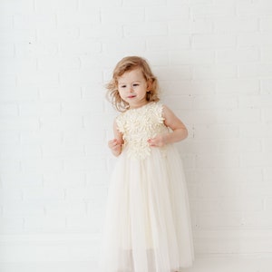 May include: A young girl wearing a white tulle dress with a floral lace bodice. She is standing in front of a white brick wall.