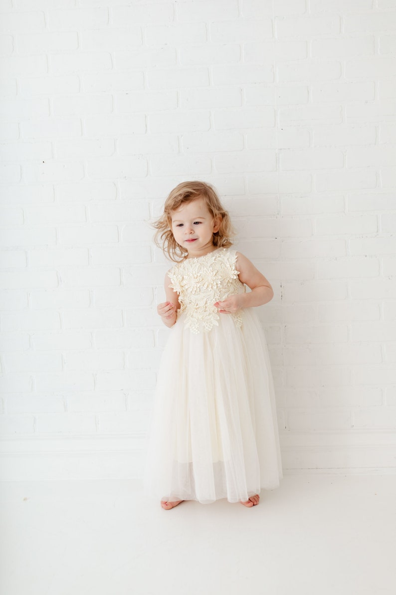 May include: A young girl wearing a white tulle dress with a floral lace bodice. She is standing in front of a white brick wall.