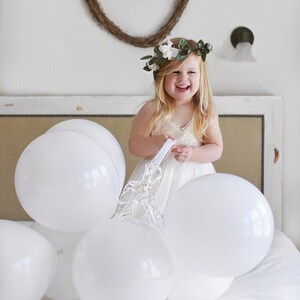 May include: A young girl wearing a white dress and a flower crown smiles while holding white balloons. The balloons are tied together with white ribbon.