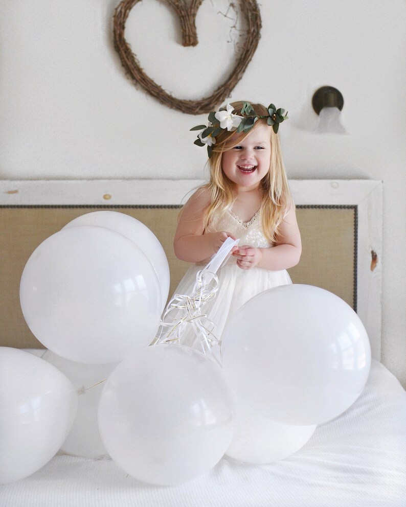 May include: A young girl wearing a white dress and a flower crown smiles while holding white balloons. The balloons are tied together with white ribbon.