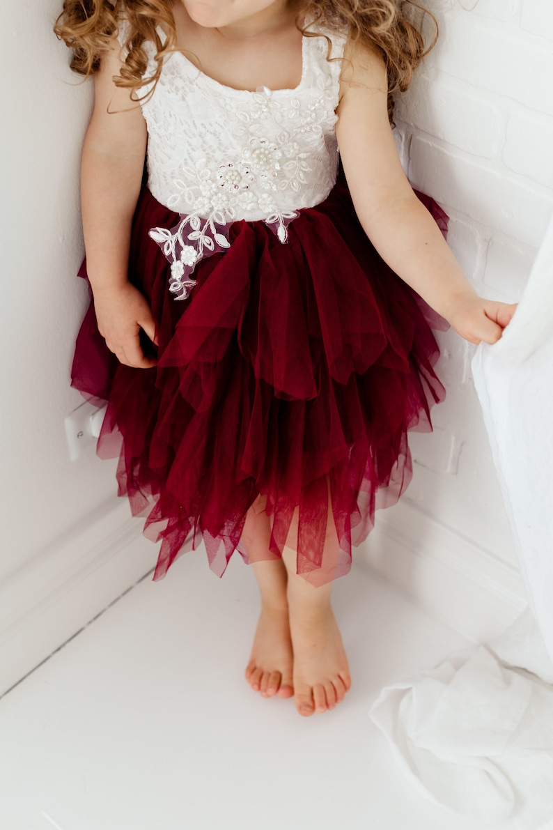 Puede incluir: Vestido sin mangas para ni&ntilde;a, con corpi&ntilde;o de encaje blanco adornado con adornos florales y falda de tul burdeos en capas. El vestido tiene un escote cuadrado y es adecuado para ocasiones especiales.