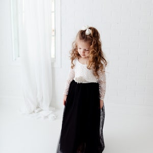 May include: A floor-length dress featuring a white lace long-sleeve bodice with a scoop neckline and a solid black tulle skirt. A decorative beaded embellishment is centered at the waistline, and a small white bow is worn in the hair.