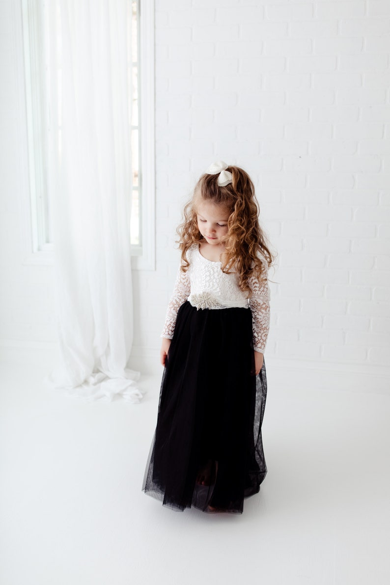May include: A floor-length dress featuring a white lace long-sleeve bodice with a scoop neckline and a solid black tulle skirt. A decorative beaded embellishment is centered at the waistline, and a small white bow is worn in the hair.