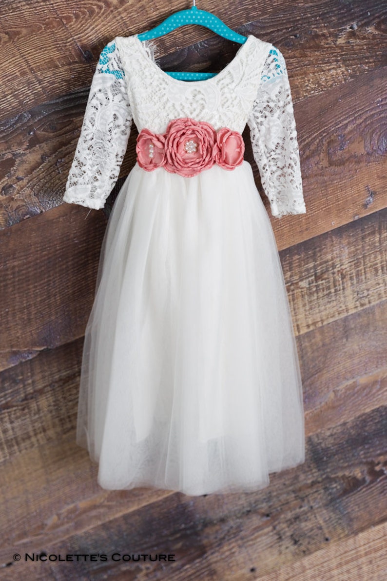 May include: A white tulle dress with a lace bodice and long sleeves. The dress has a coral flower sash.