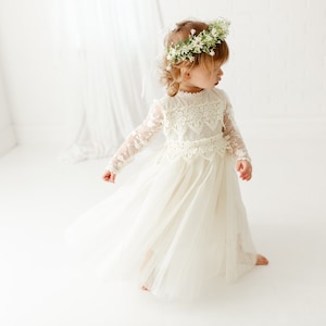 May include: A cream-colored long-sleeve dress featuring a lace bodice with a high neckline and a flowing tulle skirt. The garment is paired with a floral crown made of small white flowers and green leaves, worn atop the head.