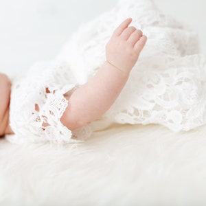 May include: A baby wearing a white lace dress and headband lies on a white fuzzy surface. The baby's arm is extended and the hand is open.