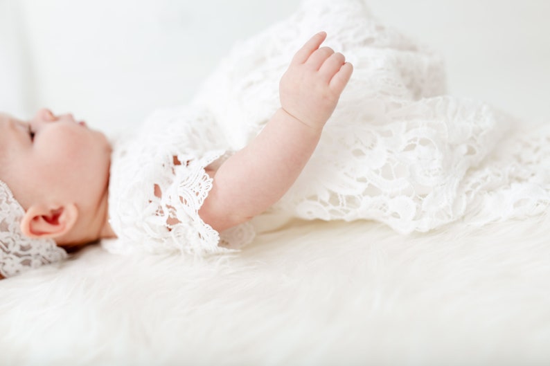 May include: A baby wearing a white lace dress and headband lies on a white fuzzy surface. The baby's arm is extended and the hand is open.