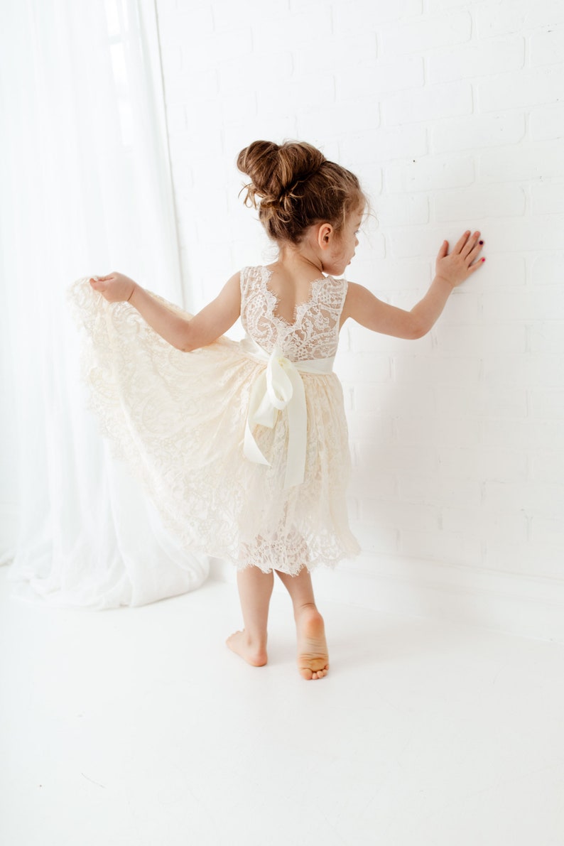 May include: A young girl wearing a white lace dress with a satin bow at the waist. The dress has a scalloped hem and a lace-up back. The girl is standing on a white surface and is looking to the side.